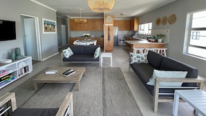A modern living room with a sofa, coffee table, and TV. A kitchen with wooden cabinets and a dining area is visible in the background. - Footprints in Kleinmond (Kleinmond)