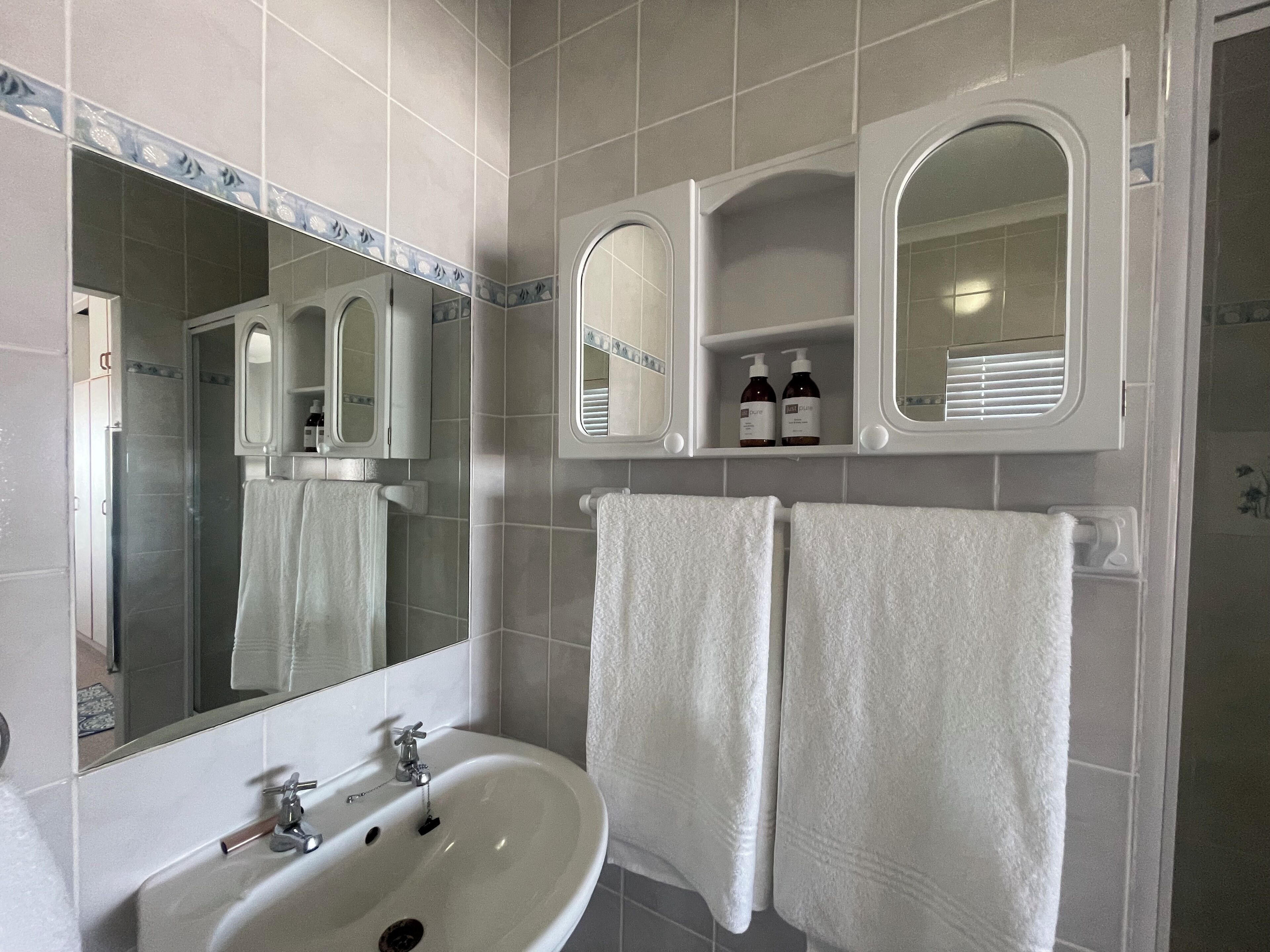 Un baño con un lavamanos blanco, dos espejos y dos toallas colgadas en la pared.