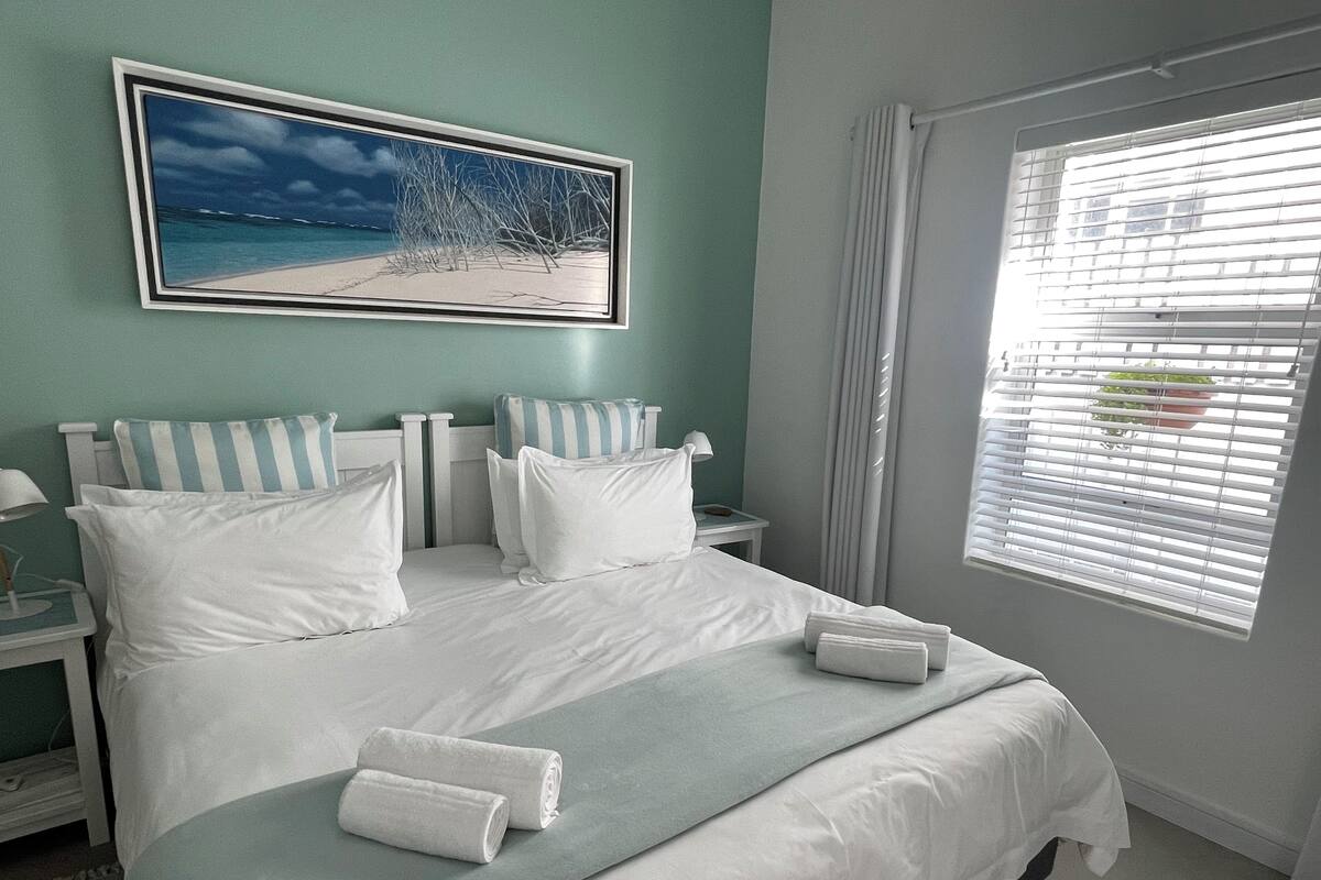 A neatly made bed with white linens and pillows, a bedside table with a lamp, and a window with blinds.