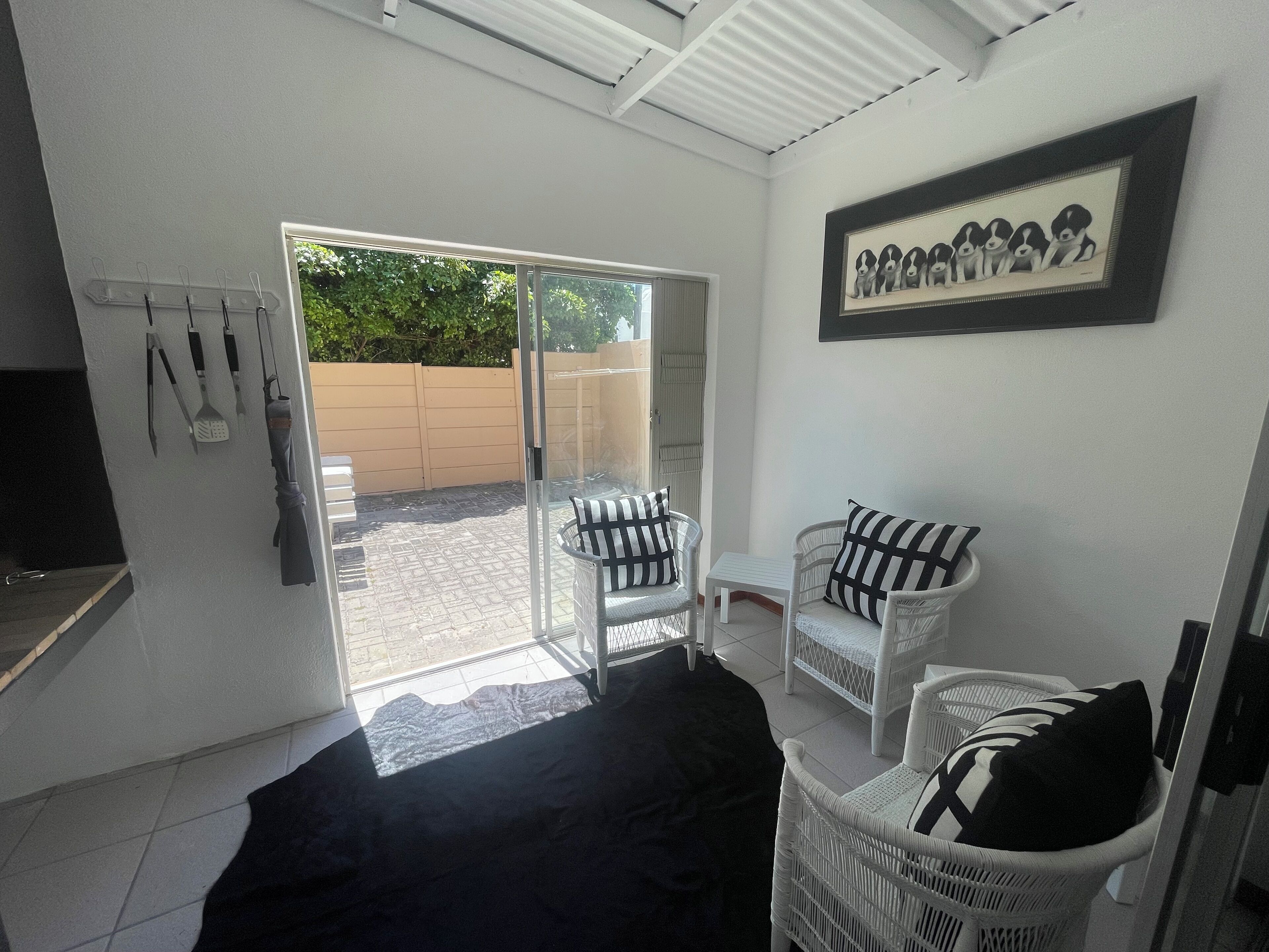 Una habitación con puerta corredera de vidrio que da a un área exterior, una silla de mimbre blanca con cojines a rayas, una pequeña mesa blanca y un cuadro con perros en la pared.