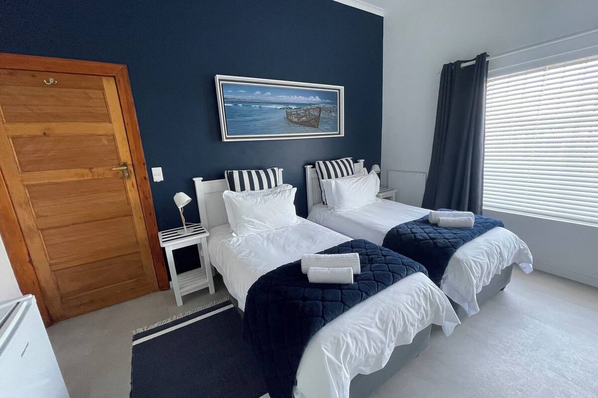 A hotel room with two beds, a wooden door, a small bedside table with a lamp, a painting of a coastal scene, and a window with blinds.