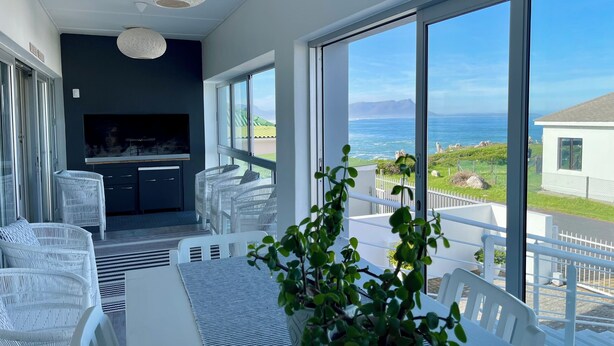 A modern dining area with a view of the ocean, white wicker furniture, and a large window. - Footprints in Kleinmond (Kleinmond)