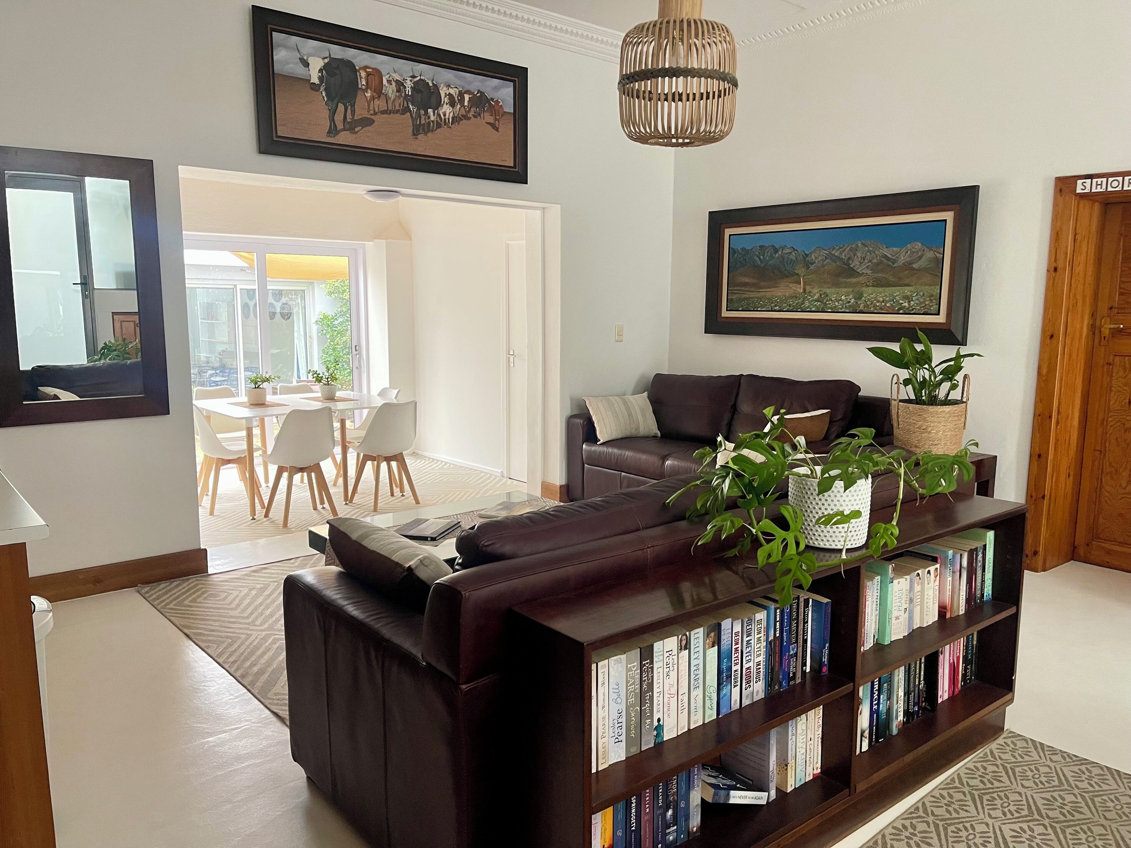 A living room with a dark brown leather sofa, a bookshelf, a dining area with a white table and chairs, and a large painting on the wall.
