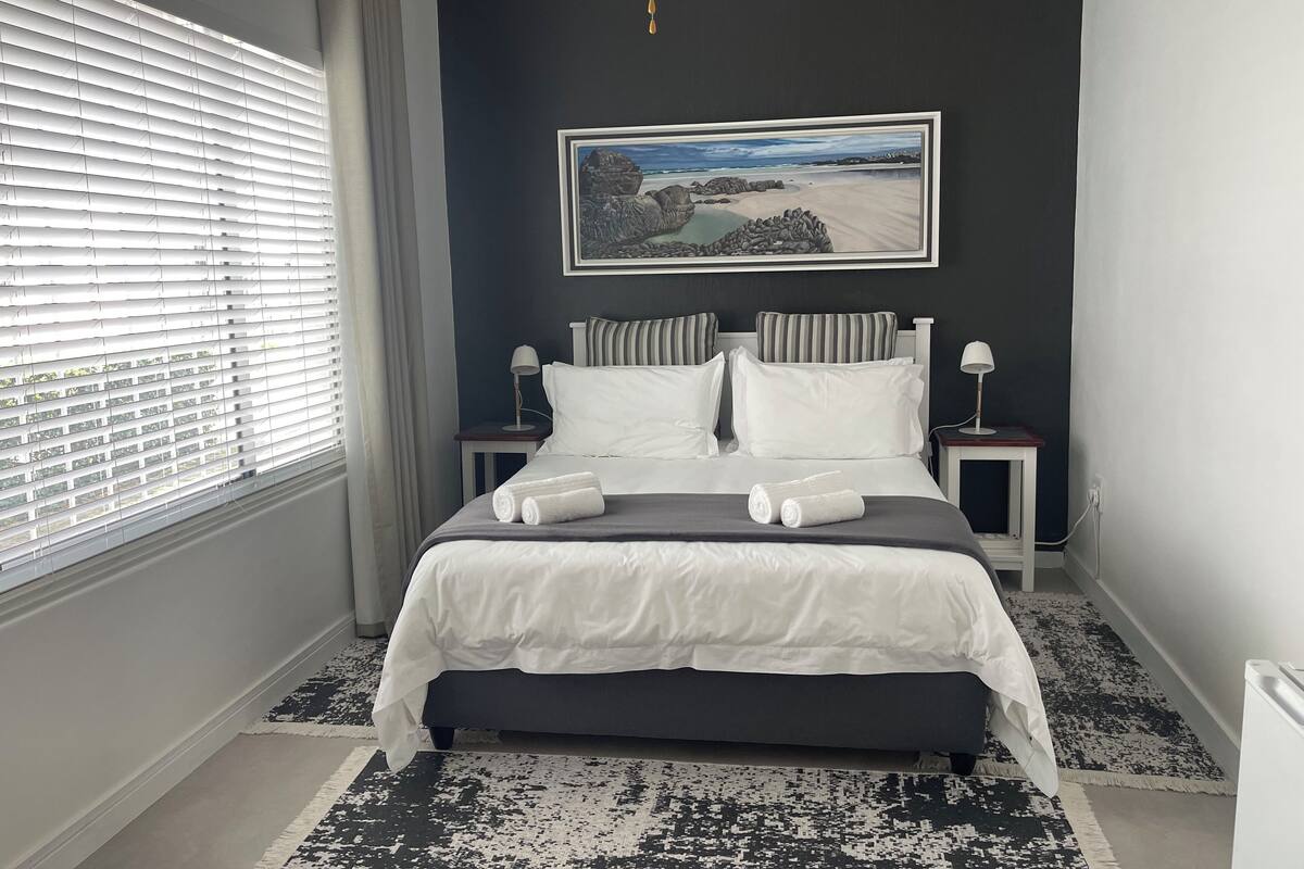 A neatly made bed with white linens and towels, a framed landscape painting on the wall, and a window with blinds.