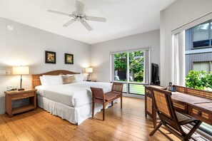 A bedroom with a bed, bedside tables, a desk, and chairs. There is a ceiling fan, a window with a view of greenery, and a television on a stand. - Oceanfront Kanaloa Condo - Pools, Hot Tubs & Sunset Views- Vrkcv (Kailua-Kona)