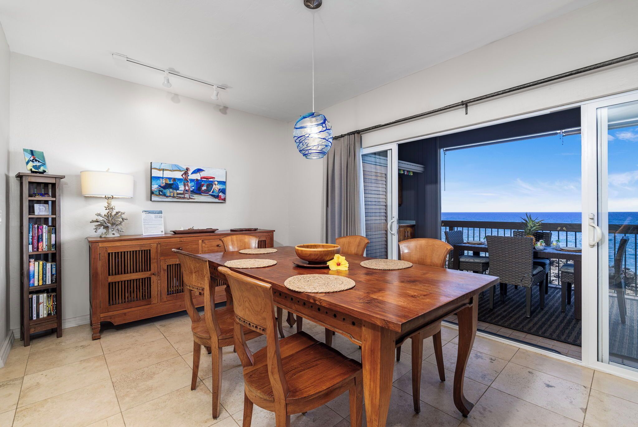 A dining area with a wooden table and chairs, a hanging light fixture, and a view of the ocean through a sliding glass door.