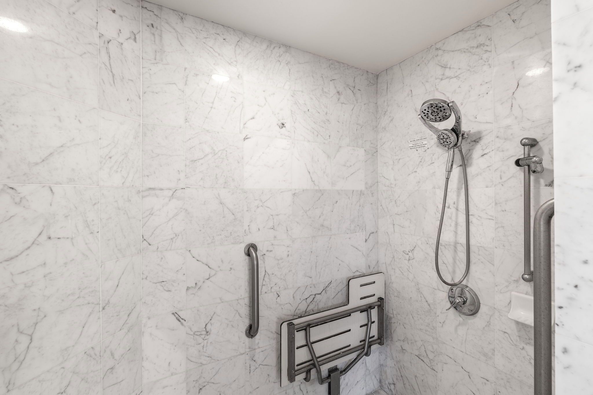 A modern bathroom with marble walls, a handheld showerhead, a wall-mounted faucet, and a grab bar for safety.