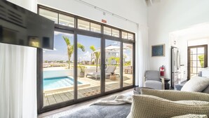A room with a large sliding glass door leading to a pool area, a sofa, a small table, and a kitchen area.