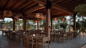An outdoor dining area with wooden tables and chairs, surrounded by palm trees and a view of the ocean.