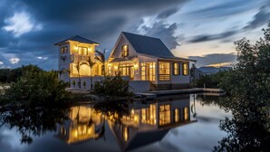 A large, two-story house with a balcony, a covered porch, and a reflection in the water.