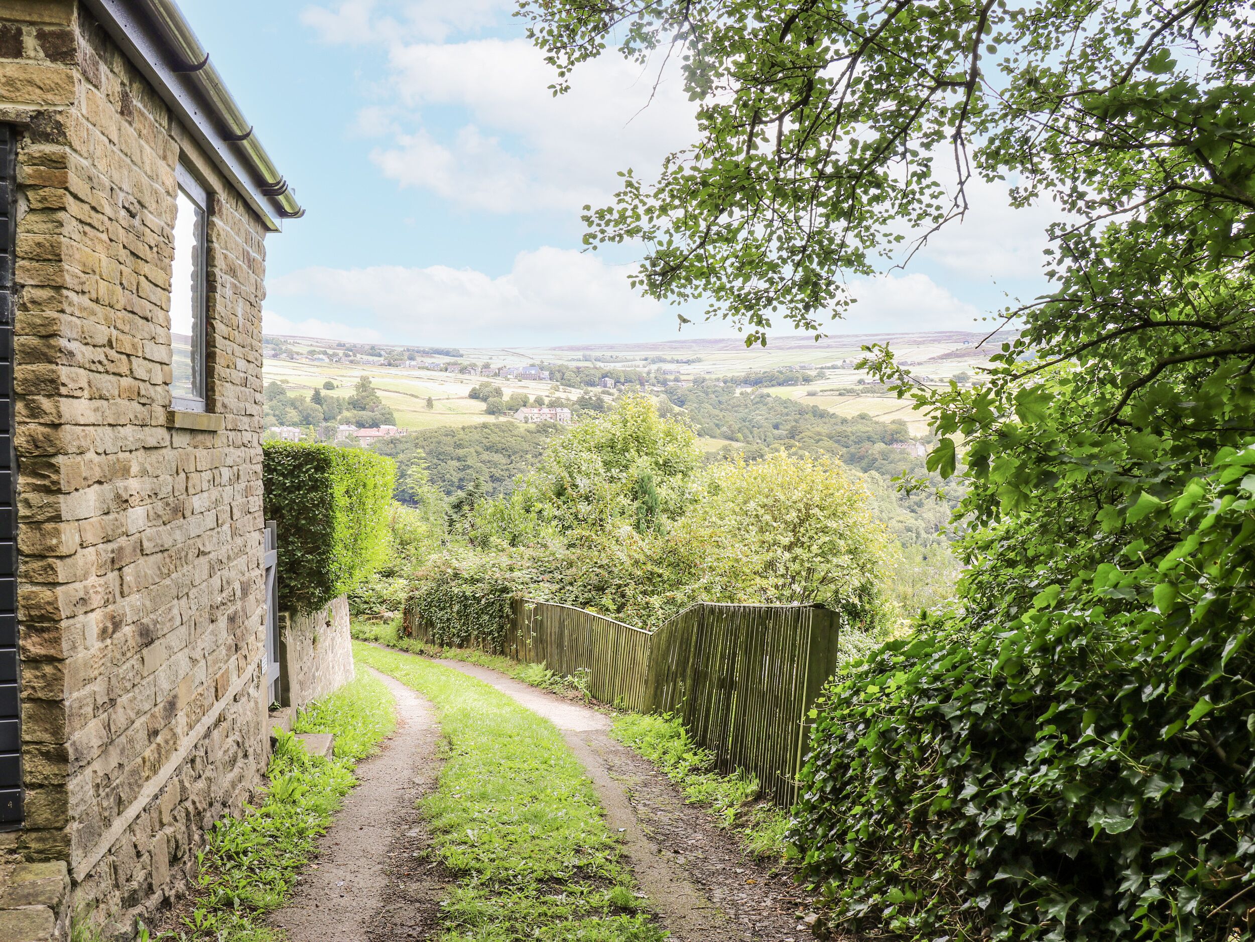 A stone building with a narrow pathway leading to a scenic view of rolling hills and a distant village.