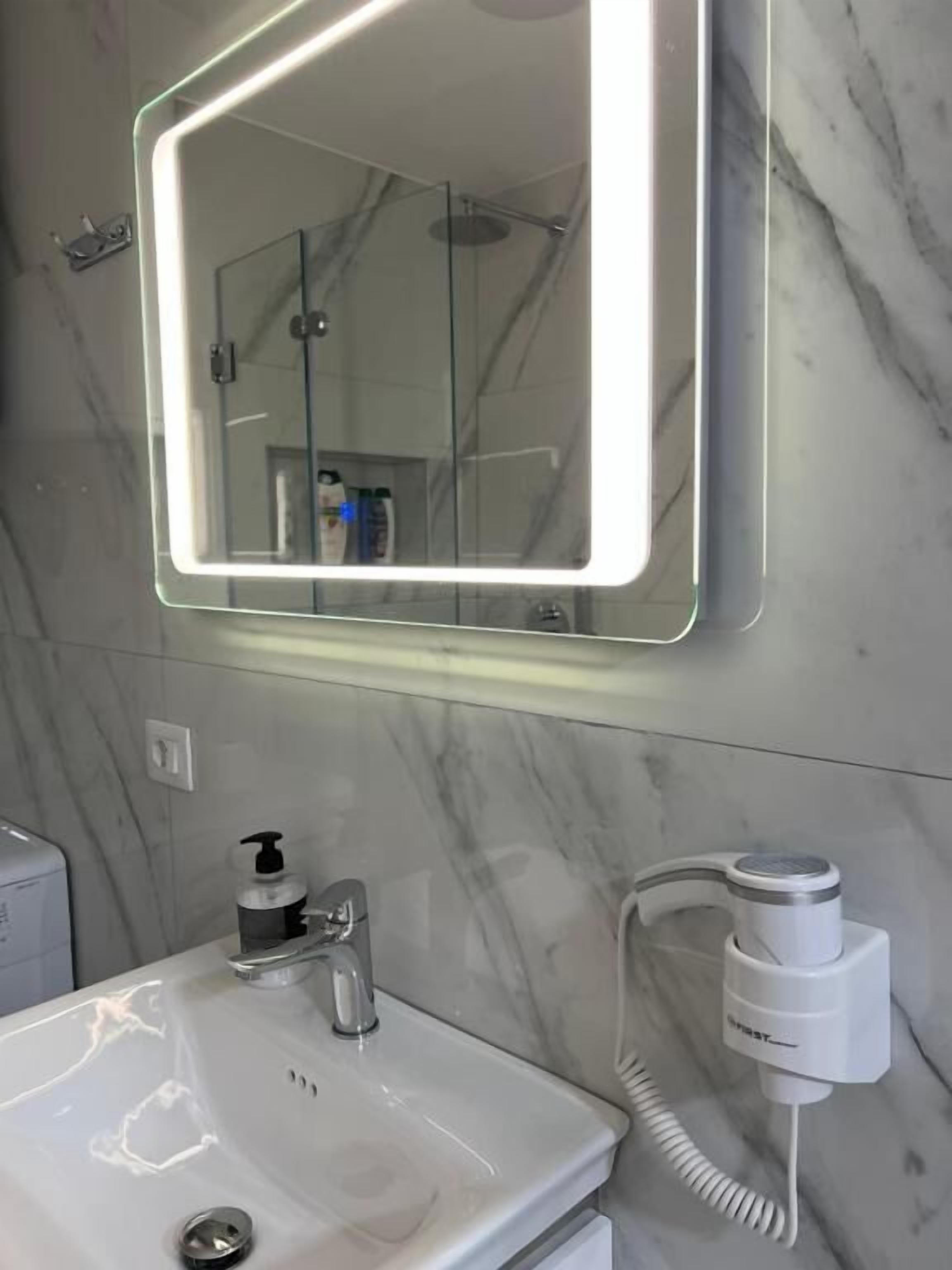 Une salle de bain avec un mur en marbre, un lavabo avec un robinet, un miroir avec un éclairage et un sèche-cheveux branché.