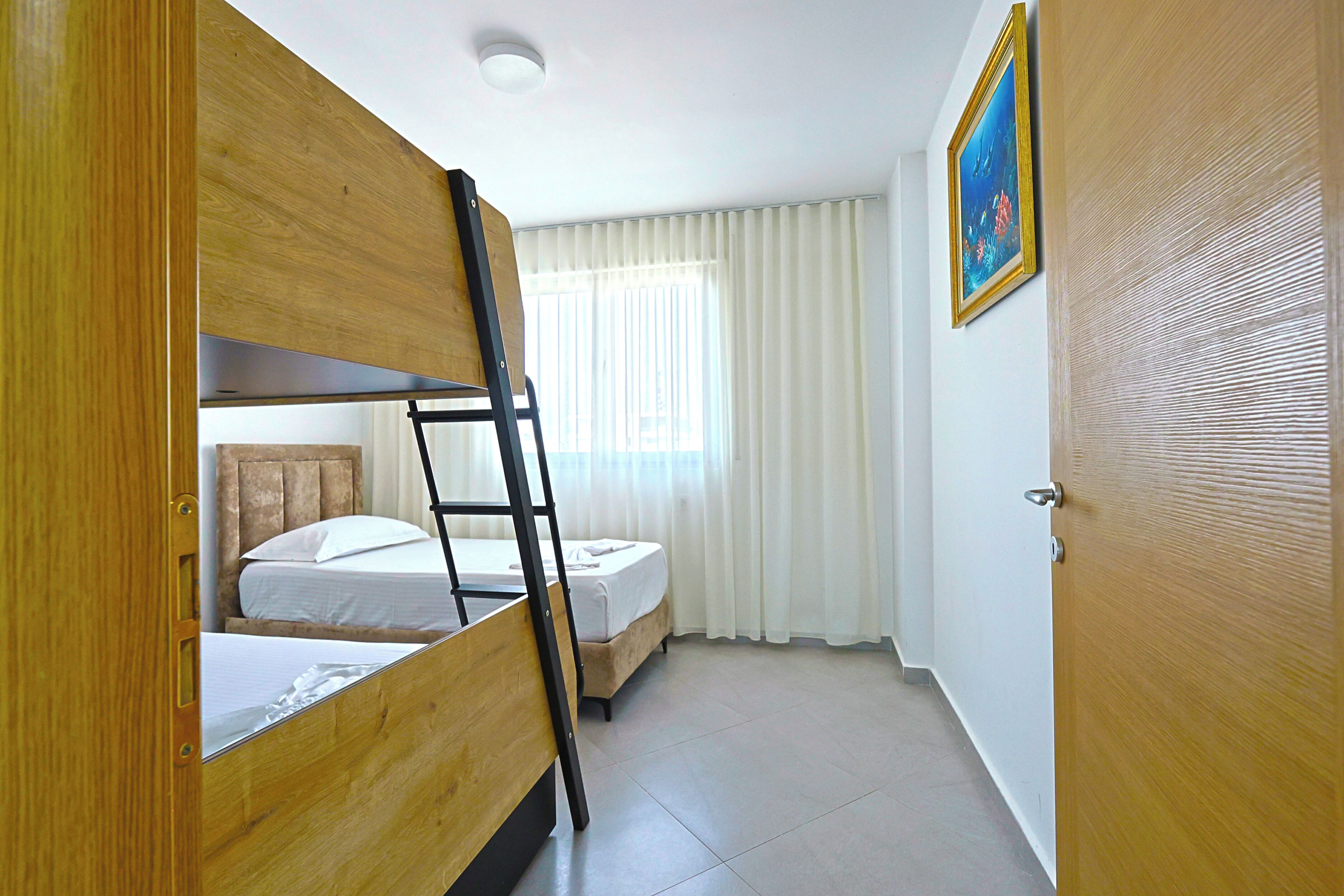 A room with bunk beds, a ladder, a bed, a window with curtains, a door, and a framed picture on the wall.