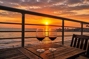 Ein Holztisch mit zwei Weingläsern auf einem Balkon mit Blick auf einen Sonnenuntergang über dem Meer.