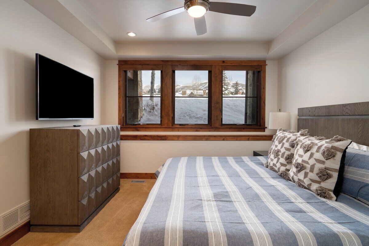 A bedroom with a large bed, a TV mounted on the wall, a window with a view of a snowy landscape, and a ceiling fan.