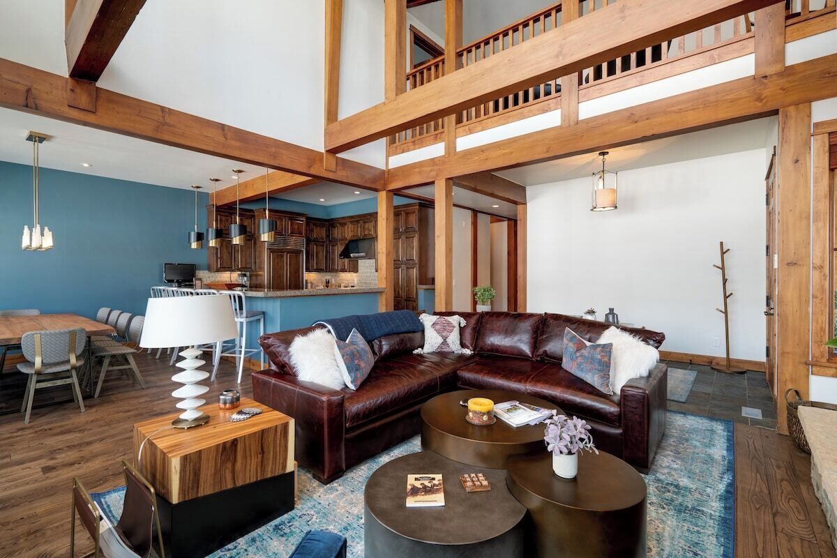 Un amplio salón con un sofá seccional de cuero oscuro, una mesa de centro de madera y un comedor con pared azul y vigas de madera.