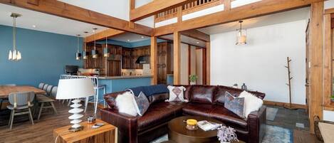 Un amplio salón con un sofá seccional de cuero oscuro, una mesa de centro de madera y un comedor con pared azul y vigas de madera.
