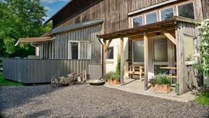 A rustic wooden building with a covered patio area, a wooden cart, and potted plants. - Vacation apartment in the Allgäu (Aichstetten)