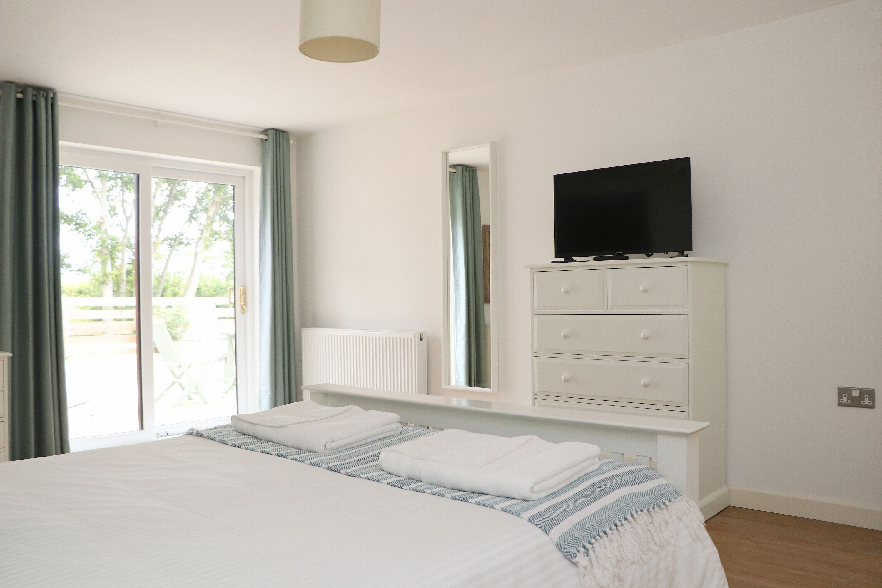Une chambre avec un grand lit, une commode sur laquelle est posée une télévision, une fenêtre avec des rideaux et une vue sur la verdure à l’extérieur.