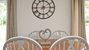 A dining area with a wooden table, white chairs, a wall-mounted clock, and a heart-shaped decoration. - Peaceful bungalow near to Constantine Bay, dog friendly with enclosed garden (Saint Merryn)
