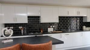 A modern kitchen with white cabinets, black tiled backsplash, and a black countertop. There are two brown chairs at a table. - Peaceful bungalow near to Constantine Bay, dog friendly with enclosed garden (Saint Merryn)