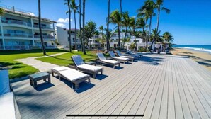 Une terrasse en bois avec des chaises longues, des palmiers et, au loin, un hôtel en bord de mer.