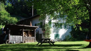 Une cabane en bois rustique, avec une table de pique-nique devant, entourée d’une végétation luxuriante et d’un bâtiment blanc aux volets verts.