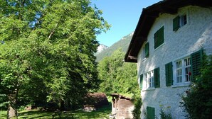 Une maison de campagne aux volets verts, entourée d’une végétation luxuriante et sous un ciel bleu azur.