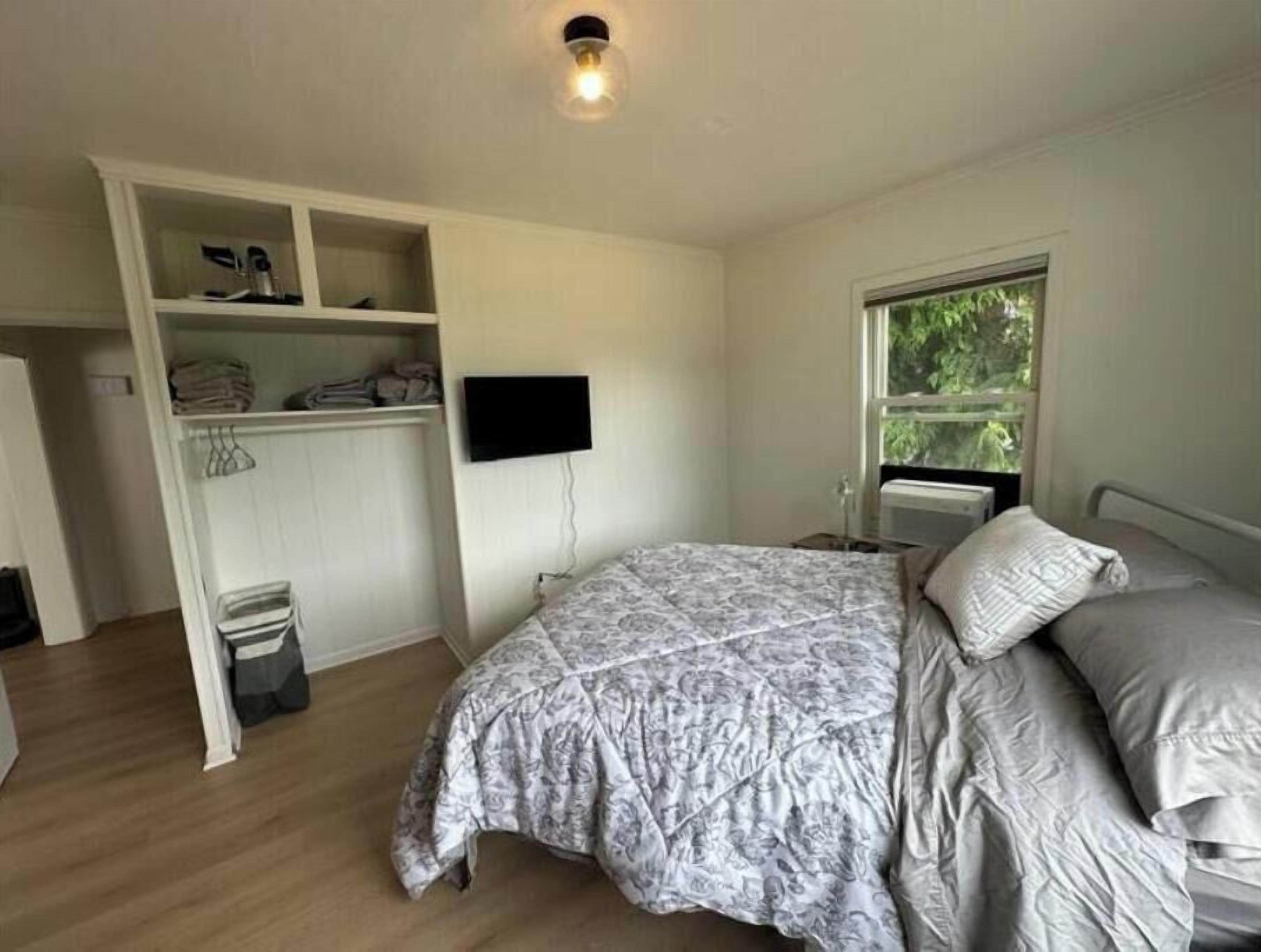 Un dormitorio con cama, televisor montado en la pared, ventana con vistas a zonas verdes y armario empotrado con estantes y espacio para colgar ropa.