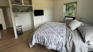 A bedroom with a bed, a TV mounted on the wall, a window with a view of greenery, and a built-in wardrobe with shelves and hanging space.
