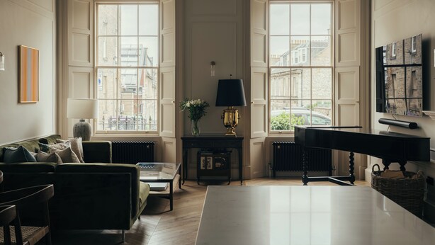 A spacious living room with a piano, a sofa, a coffee table, and a large window providing a view of the city.
