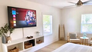 A modern hotel room with a large bed, a mounted TV displaying a movie, a white console table, and a large window. - Pet-Friendly Estero Spacious House • Near FGCU, Beaches, RSW Airport & Hertz (Fort Myers)