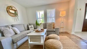 A modern living room with a grey sofa, a white coffee table, and two round poufs. There are two windows with blinds, a floor lamp, and a wall-mounted shelf with a plant. - Pet-Friendly Estero Spacious House • Near FGCU, Beaches, RSW Airport & Hertz (Fort Myers)