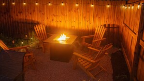 Una terraza de madera con una mesa de pozo de fuego rodeada de sillas Adirondack, luces colgantes y una pequeña planta en maceta.