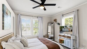Ein Schlafzimmer mit einem Bett, einem Deckenventilator, einem Fernseher auf einer weißen Konsole und einem Fenster mit Vorhängen.