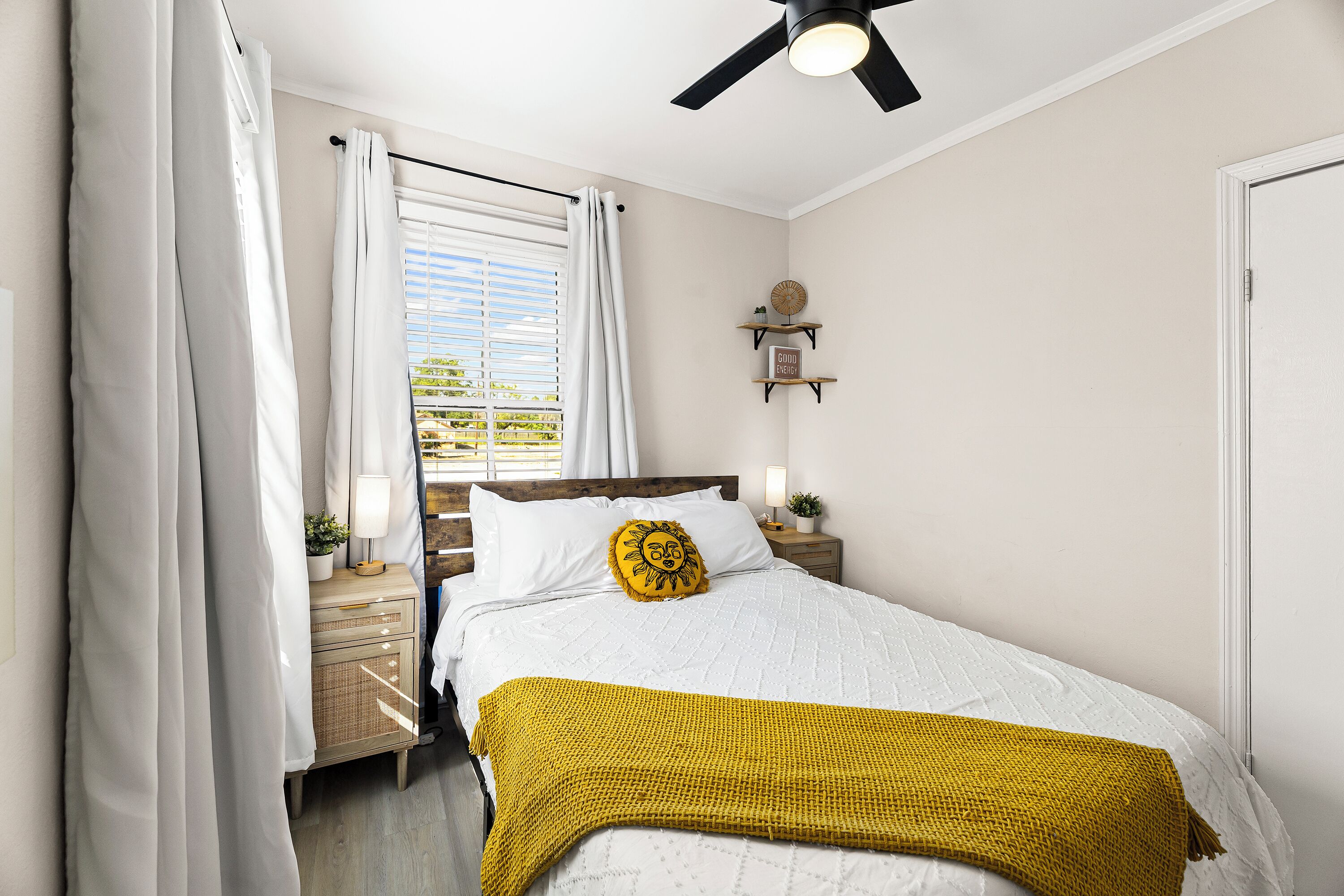 Ein Schlafzimmer mit einem Bett, einem Nachttisch, einem Deckenventilator, einem Fenster mit Vorhängen und einem an der Wand befestigten Regal mit einer Uhr und einem Schild.
