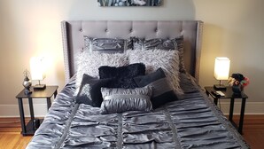 Una cama bien tendida con un edredón de satén gris y varios almohadones, un cabecero capitonado y un cuadro floral sobre ella.