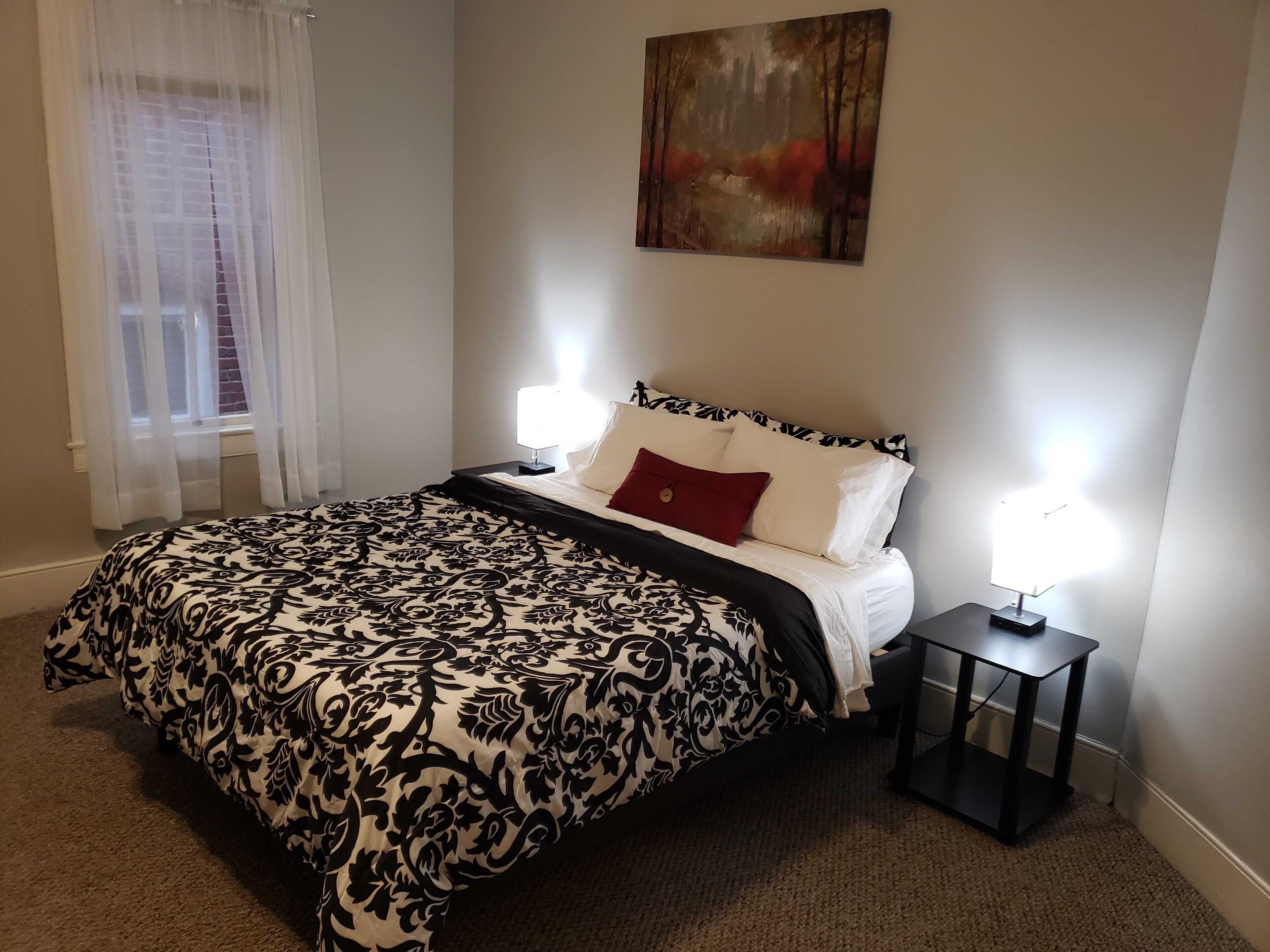 A neatly arranged bedroom with a large bed, two bedside lamps, a painting on the wall, and a window with sheer curtains.