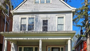 Una casa de dos pisos con porche delantero, chimenea de ladrillo y un pequeño jardín frontal.