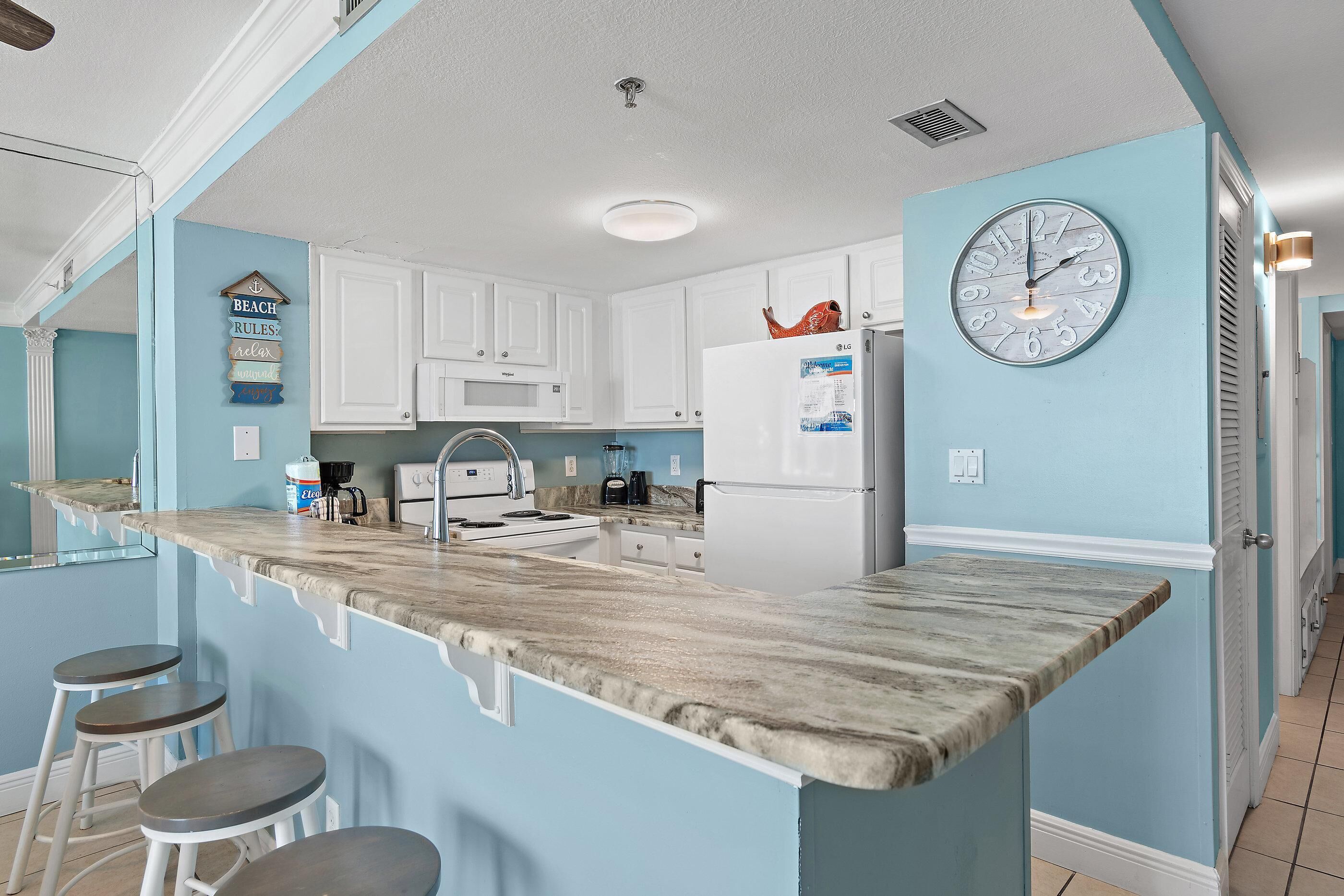 A kitchen with a marble countertop, white cabinets, a clock, and a sign that says "BEACH RULES."