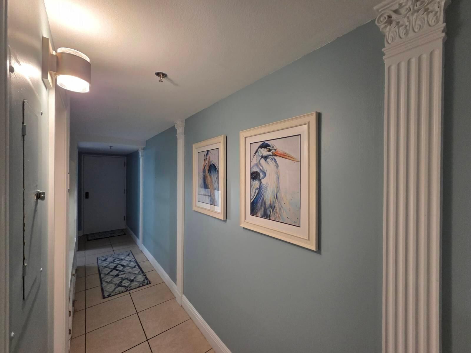 Un couloir aux murs bleu clair, orné de deux tableaux encadrés, un tapis à motifs et une colonne blanche.
