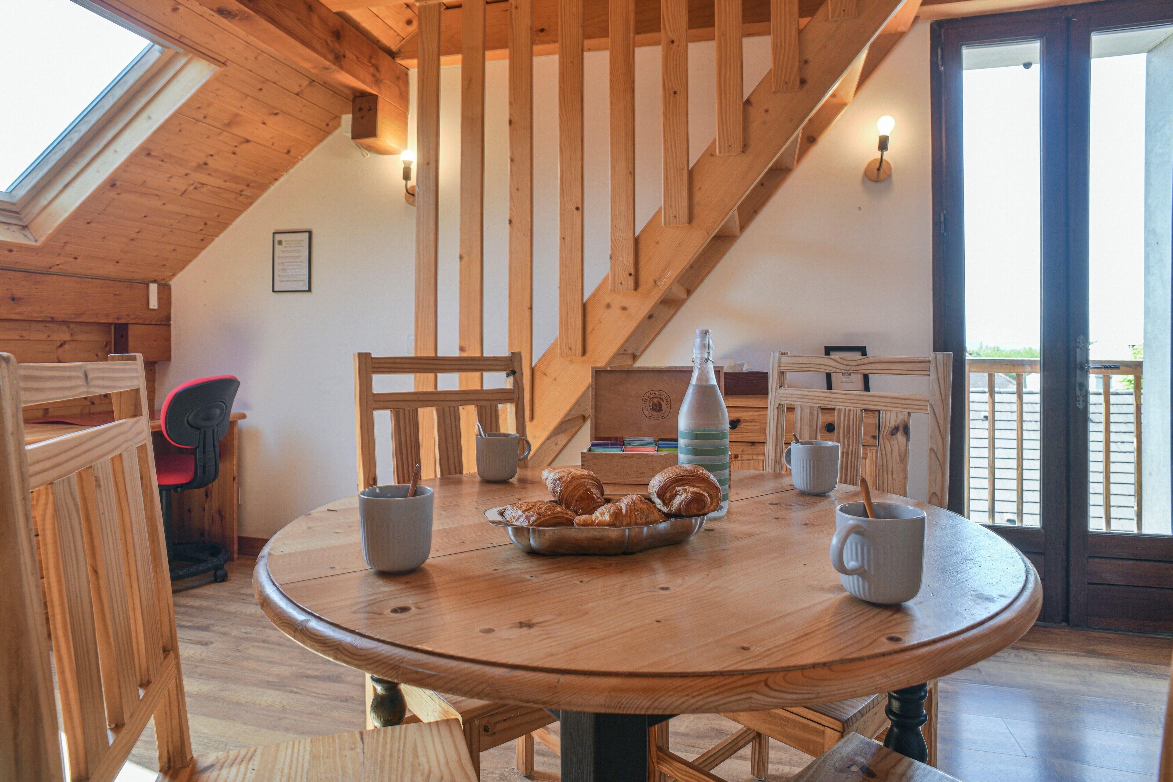 A wooden dining table with chairs, a tray of croissants, and a bottle. A staircase with wooden railings is visible in the background.