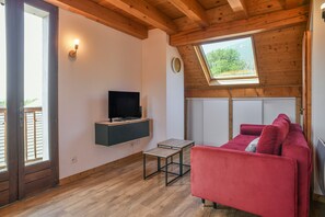 Una habitación con techo de madera, un sofá rojo, un televisor sobre un soporte negro, una mesita y una ventana con vistas a zonas verdes.