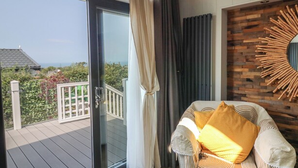 A balcony with a view of the sea, a wicker chair with a cushion, and a wooden wall with a sunburst mirror. - Coogee View (New Quay)