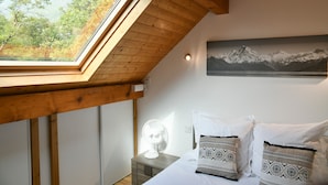 A bedroom with a skylight, a bed with patterned pillows, a nightstand with a fan, and a mountain landscape painting on the wall. - Chez Fred 201 – Parking, Wi-Fi, Near Savoie College (Saint-Pierre-d'Albigny)