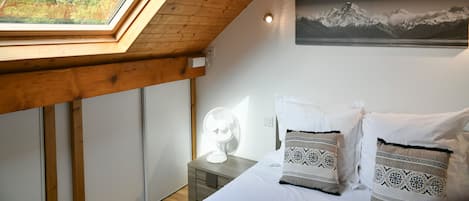 A bedroom with a skylight, a bed with patterned pillows, a nightstand with a fan, and a mountain landscape painting on the wall.