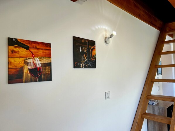 Ein Zimmer mit Holzbalkendecke, weißer Wand und zwei Kunstwerken, die Wein und ein Fass zeigen.