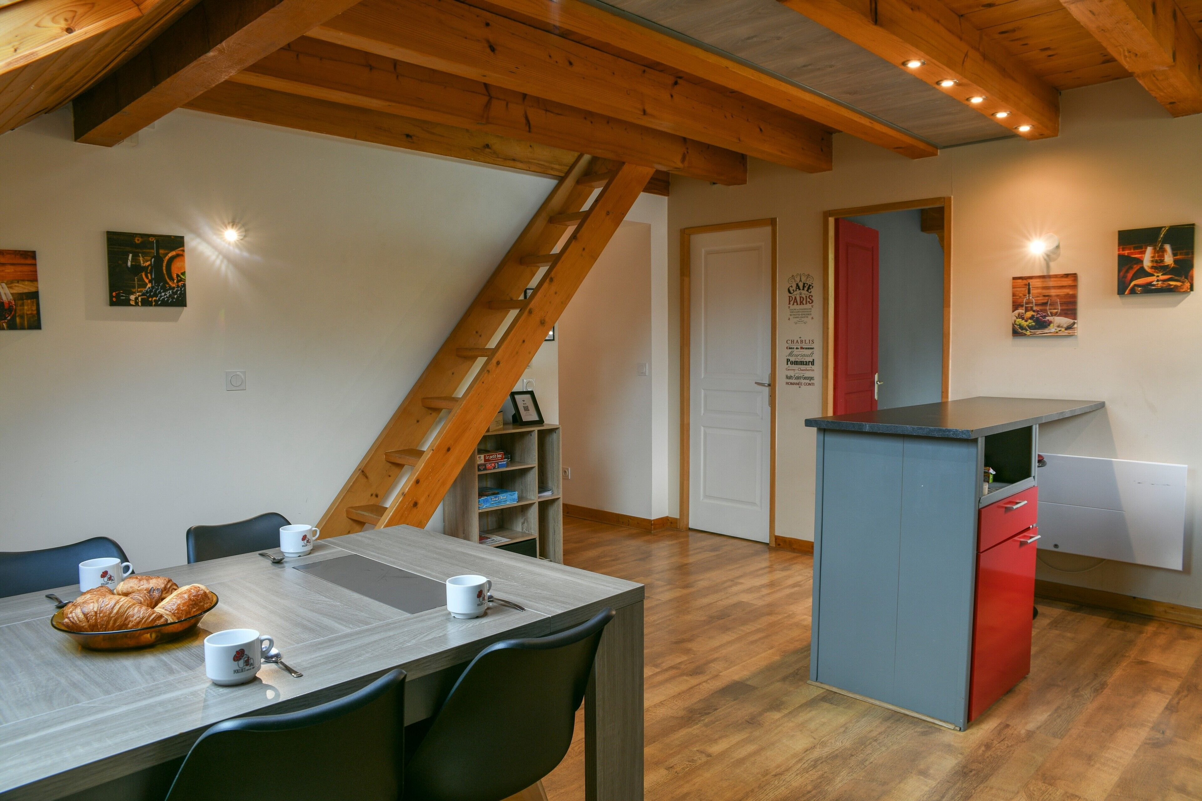 Un coin repas avec une table dressée pour deux personnes, une échelle et un espace cuisine équipé d’un réfrigérateur.