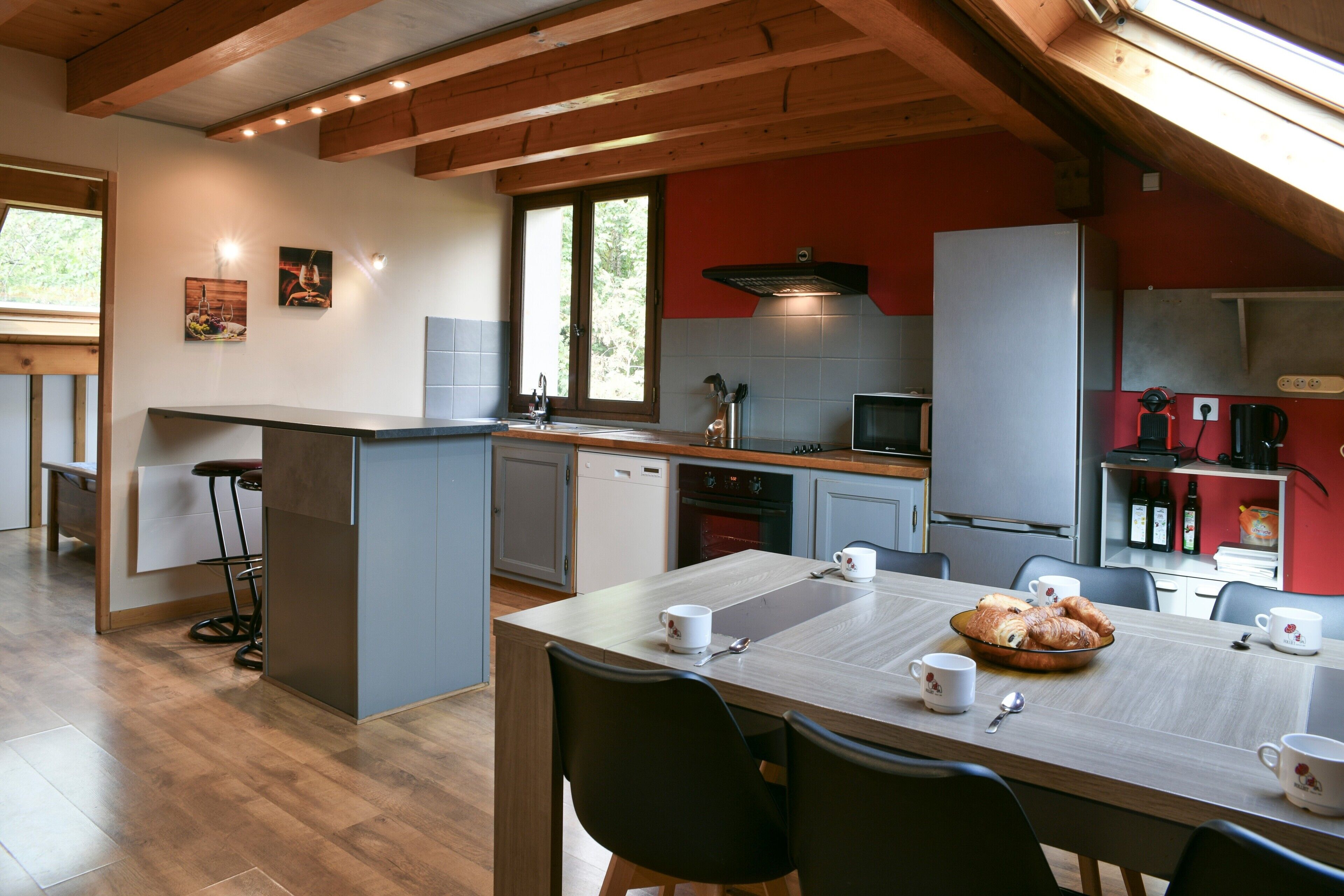 Une cuisine moderne avec des poutres en bois, un coin repas avec des chaises, un espace pour prendre le petit-déjeuner et un mur d’accent rouge.