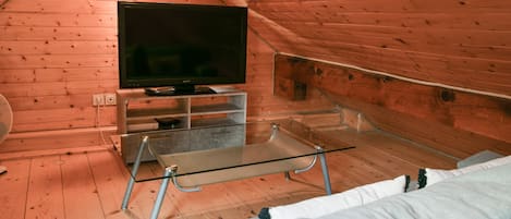 A cozy attic room with a television on a glass table, wooden walls, and a sofa.